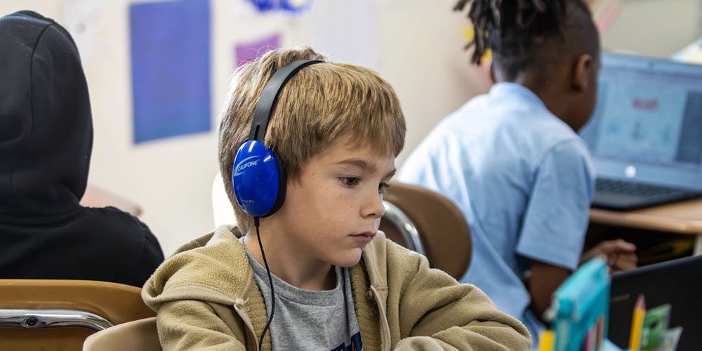 Student wearing blue headphones working at a classroom desk.