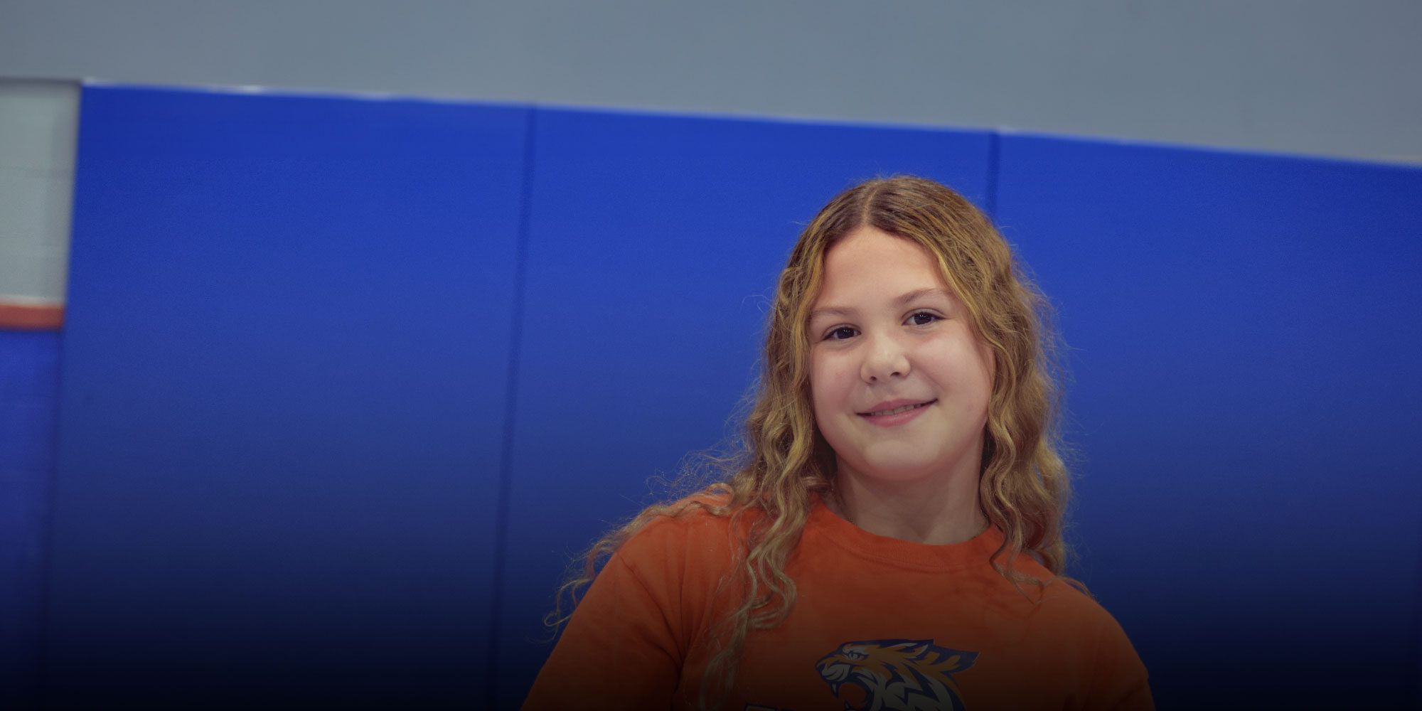 Smiling student in an orange tiger logo shirt against a blue wall.