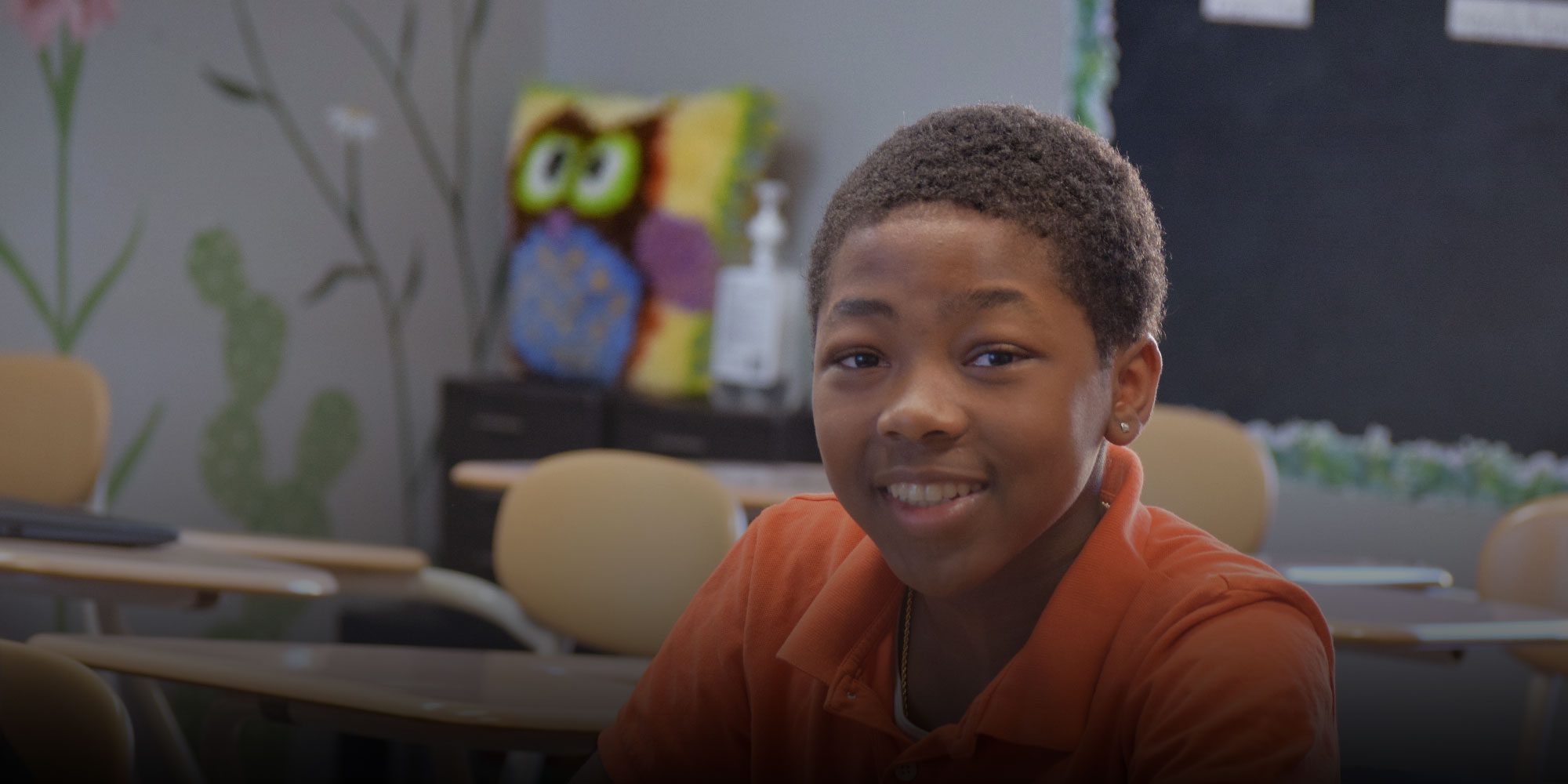 Smiling student in an orange polo shirt at a classroom desk.